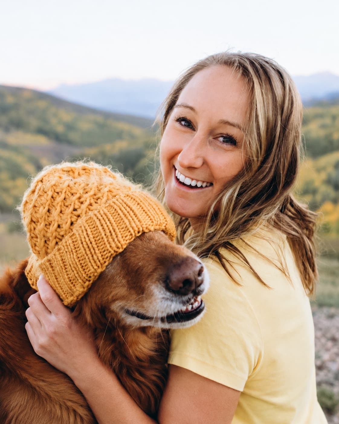 A smiling customer hugging her dog, who is wearing a yellow knit beanie